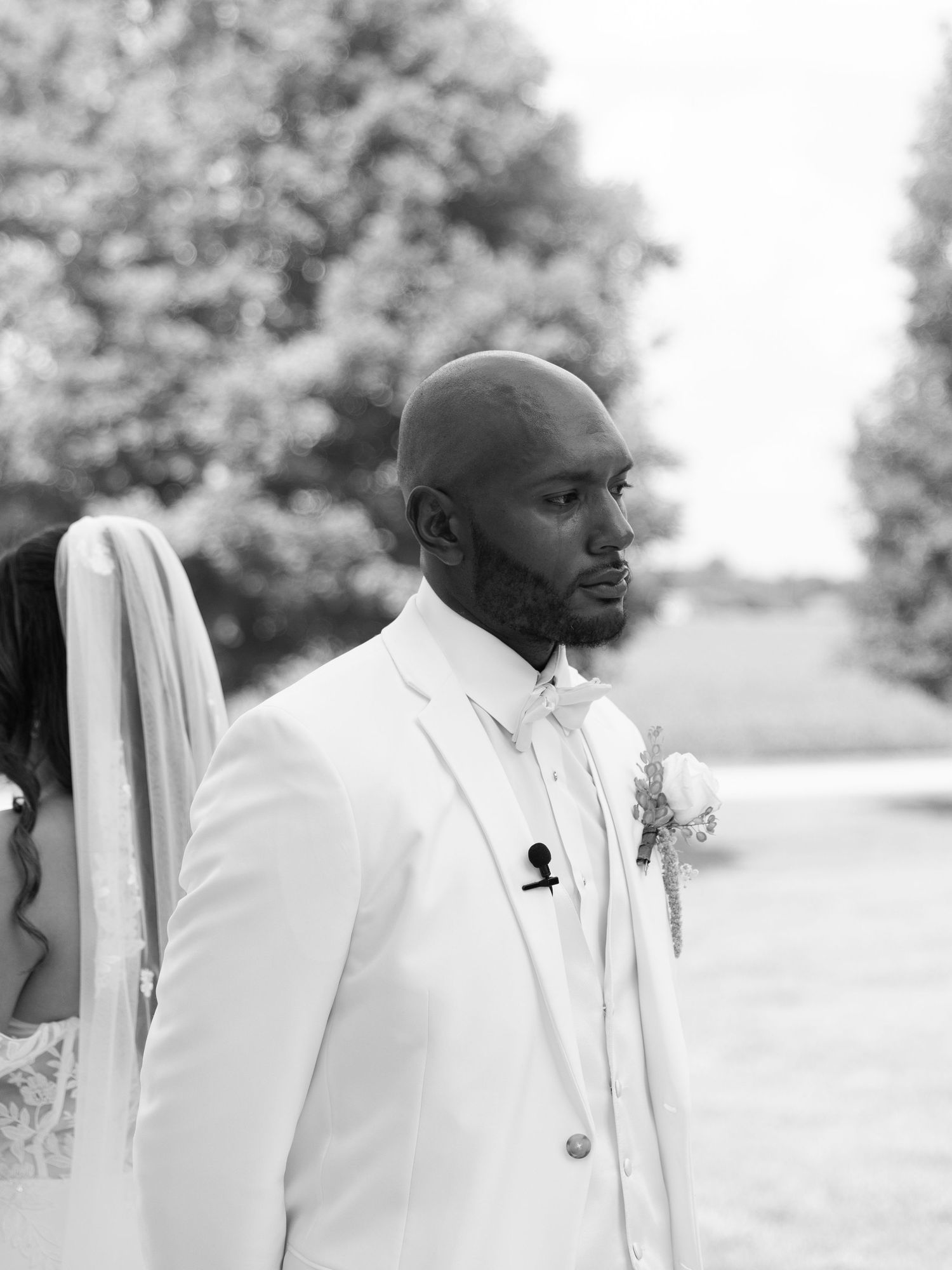 Black and white photo of a man helping another man in a white suit jacket get dressed for a wedding.