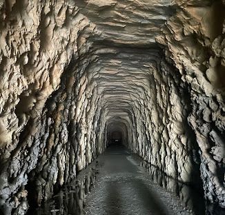 A dark rocky tunnel carved through stone stretches into the distance with rough textured walls illuminated by a distant light.