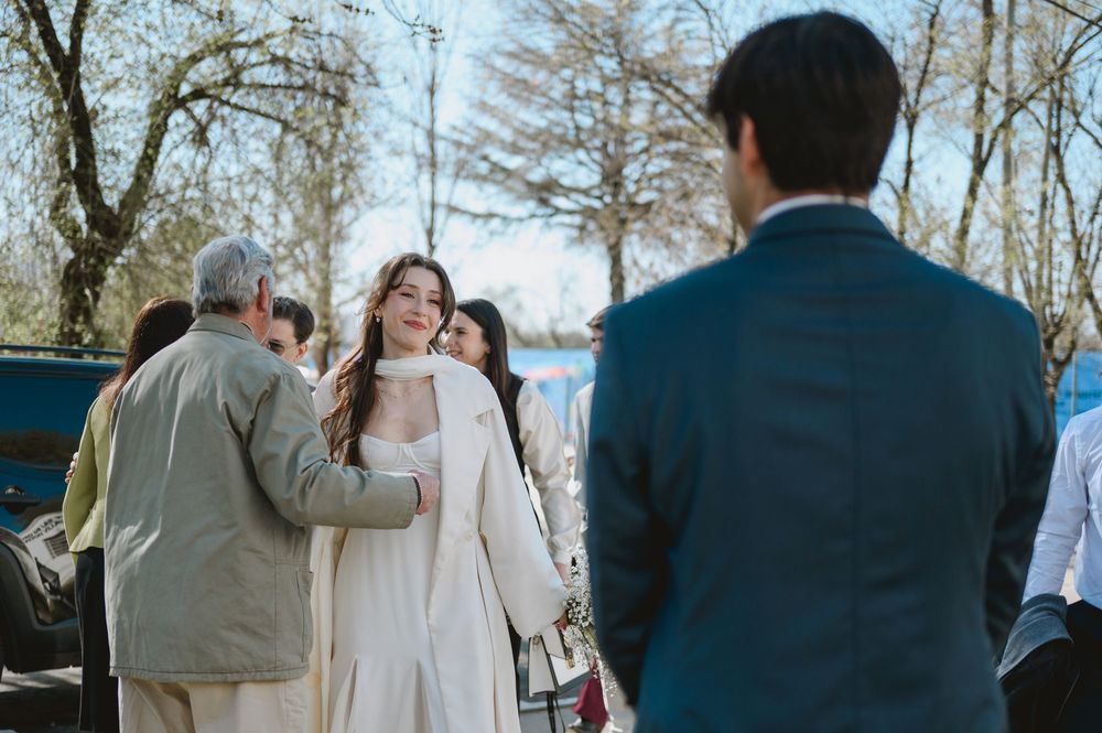 Bride in white coat looking toward groom while older guest stands nearby at outdoor wedding.