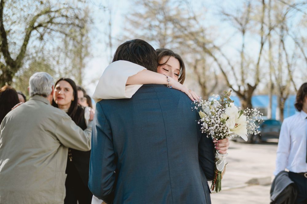 Bride and groom sharing a warm embrace outdoors after their wedding ceremony while guests celebrate nearby.