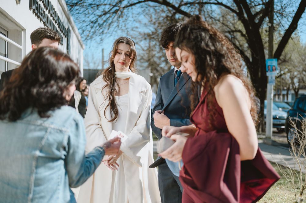 Bride in a white coat greets guests outside on a sunny day during a wedding celebration.