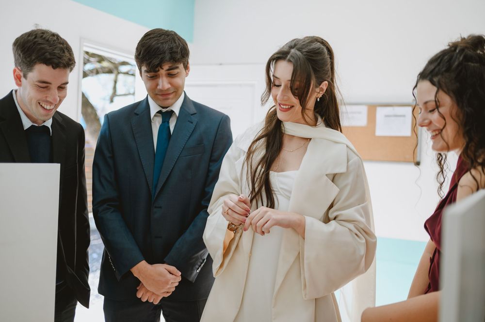 Bride in white coat laughs with groom and guests while examining wedding rings indoors before ceremony.