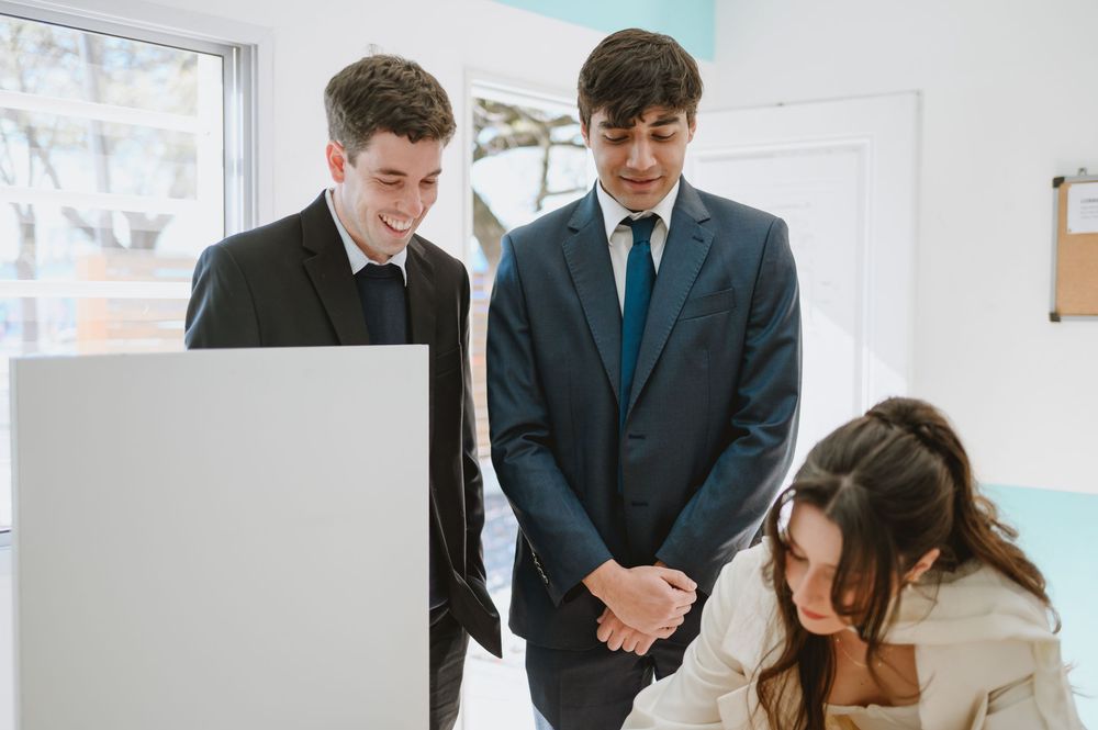 Two men in suits smile while watching a woman sign documents at a white podium.