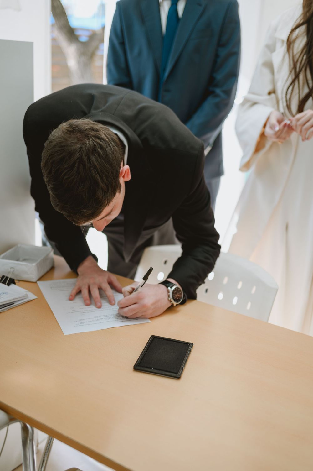 Groom in black suit signing wedding documents at a wooden table during a civil ceremony.