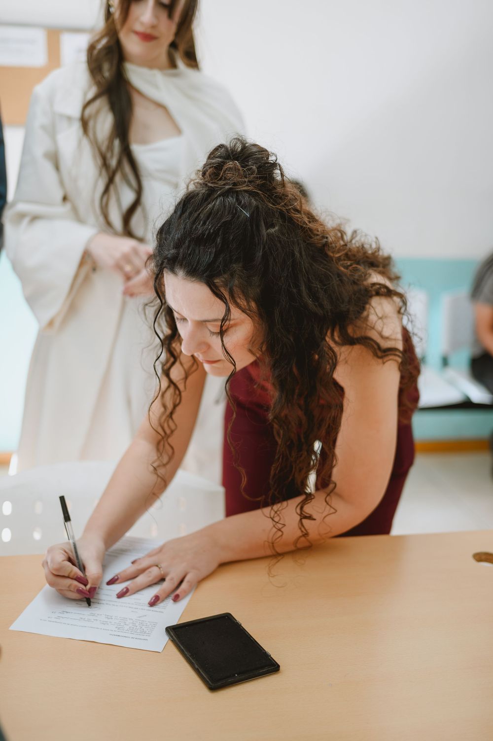 Woman with curly dark hair in burgundy dress signing documents at a wooden table during a wedding ceremony.