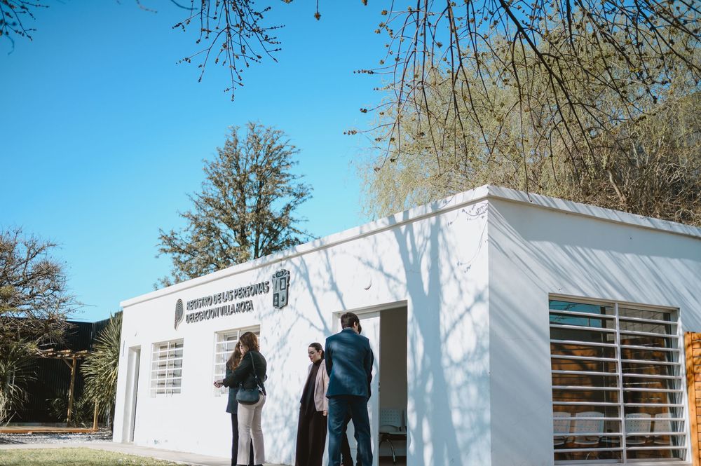 People standing outside a modern white building with a sign in bright sunlight surrounded by trees.