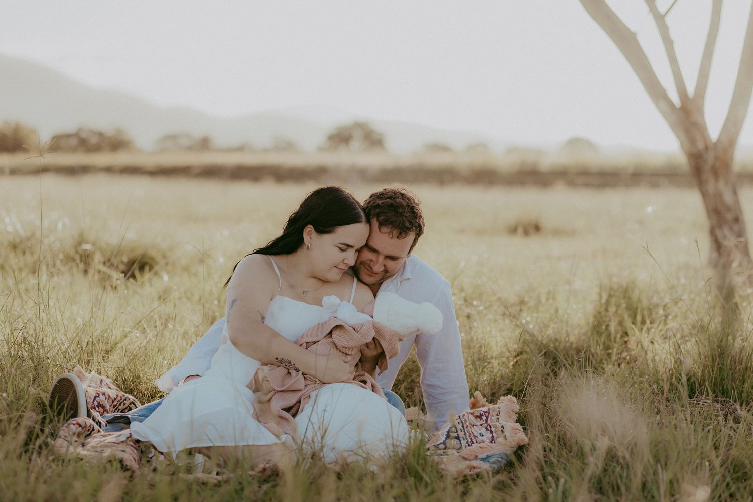 Couple sitting in golden grass field at sunset, sharing tender moment with newborn baby.