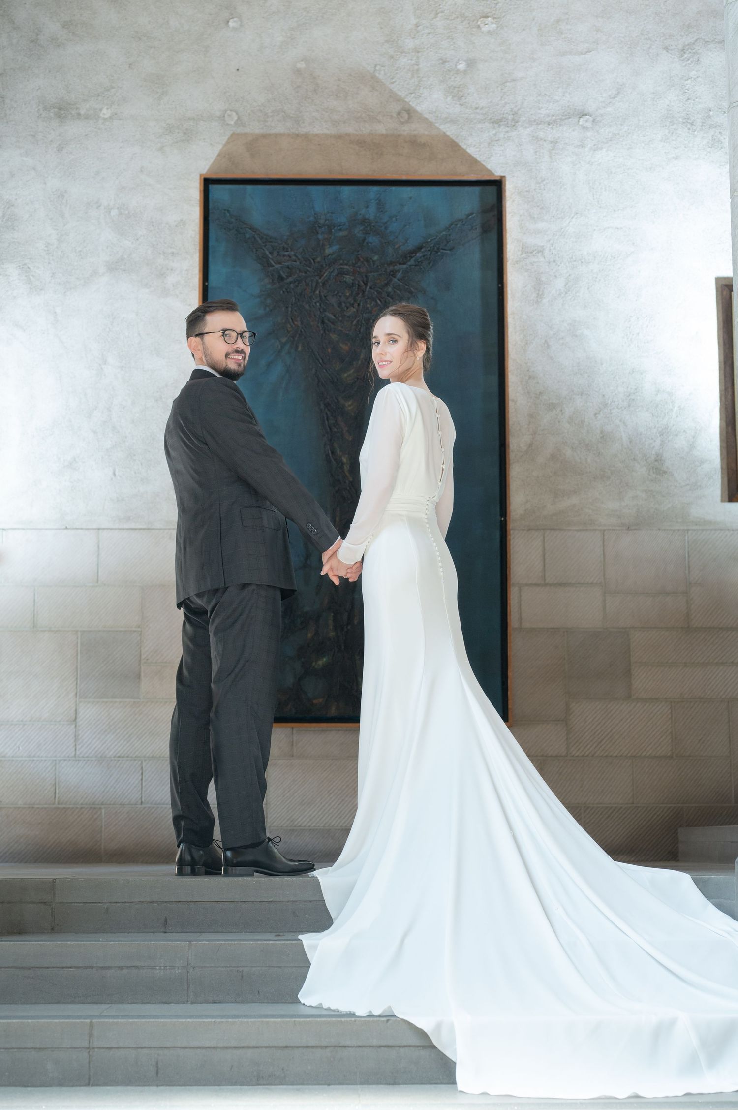 Bride and groom holding hands in elegant wedding attire, posing before a dark artwork on stone steps.