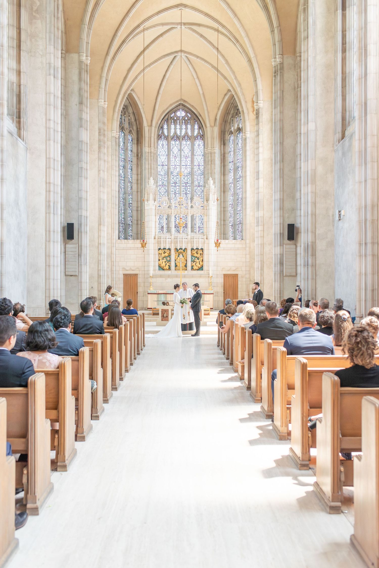 Wedding ceremony in a grand Gothic church with tall arched windows, wooden pews filled with guests, and couple at the altar.