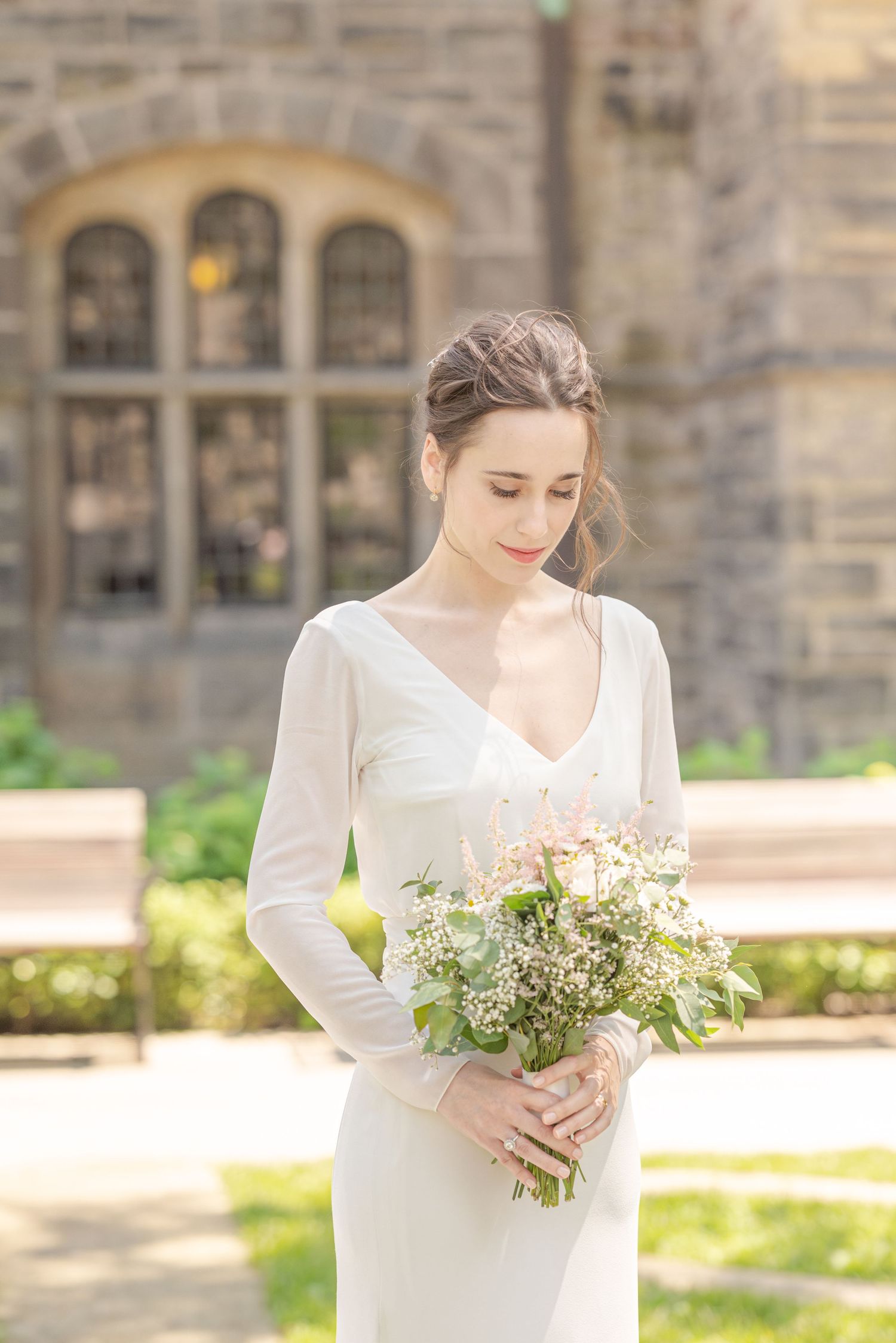 Bride in white long-sleeve gown holding wildflower bouquet, standing before stone building outdoors.