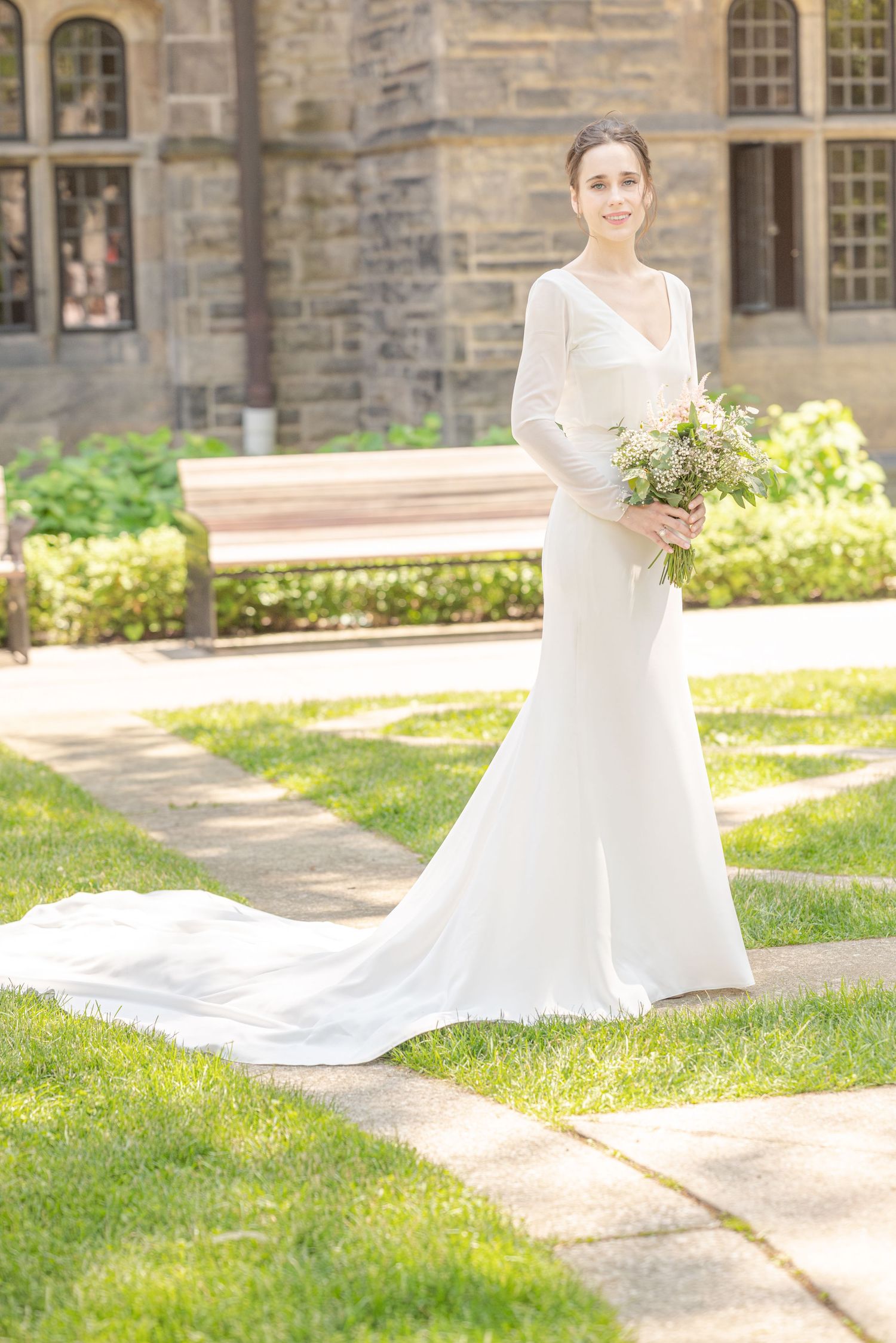 Bride in elegant long-sleeve white gown with train holds bouquet outdoors near stone building on sunny day.
