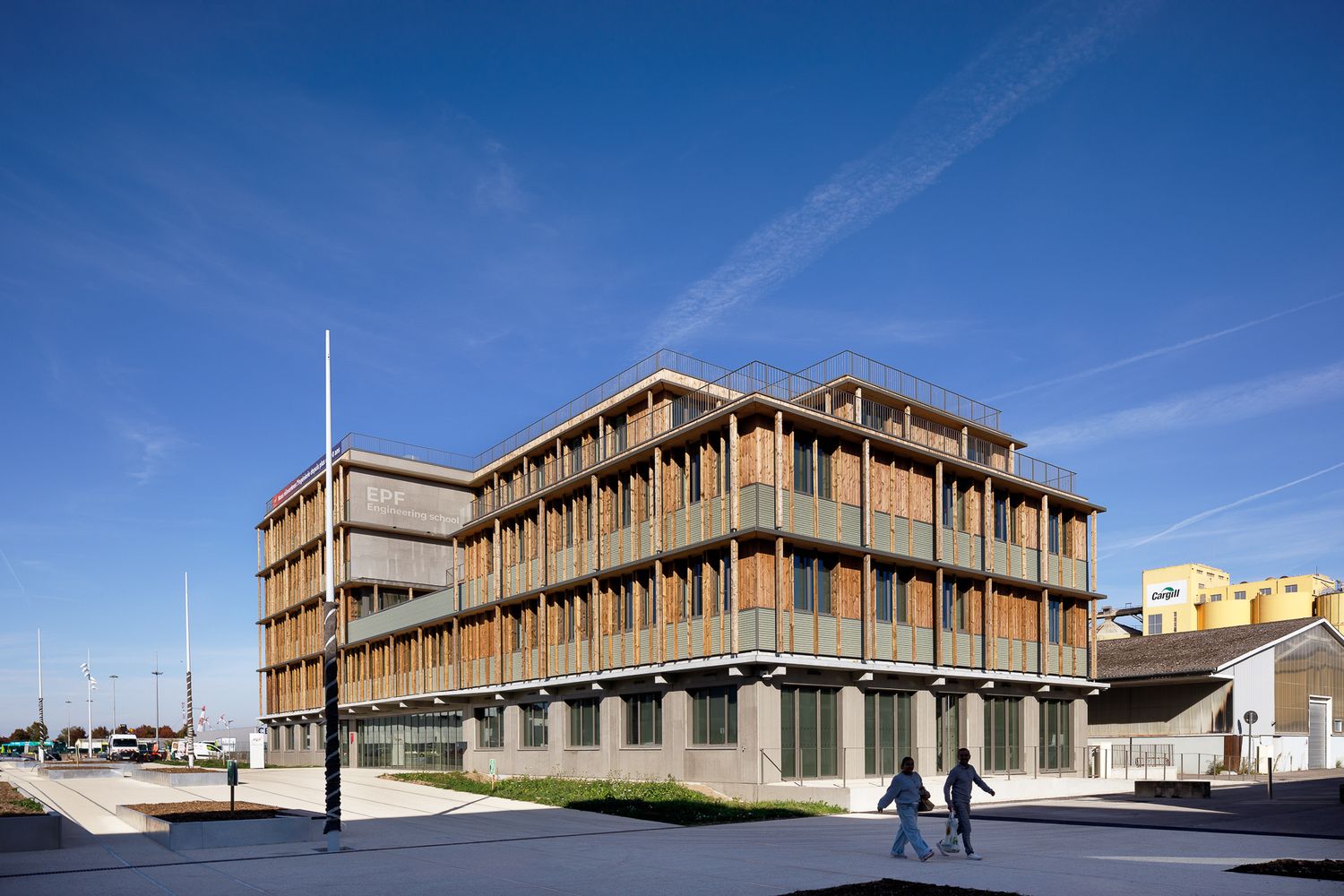 Bâtiment moderne de quatre étages avec une façade dotée de brise-soleil en bois et un revêtement végétalisé, sous un ciel bleu.