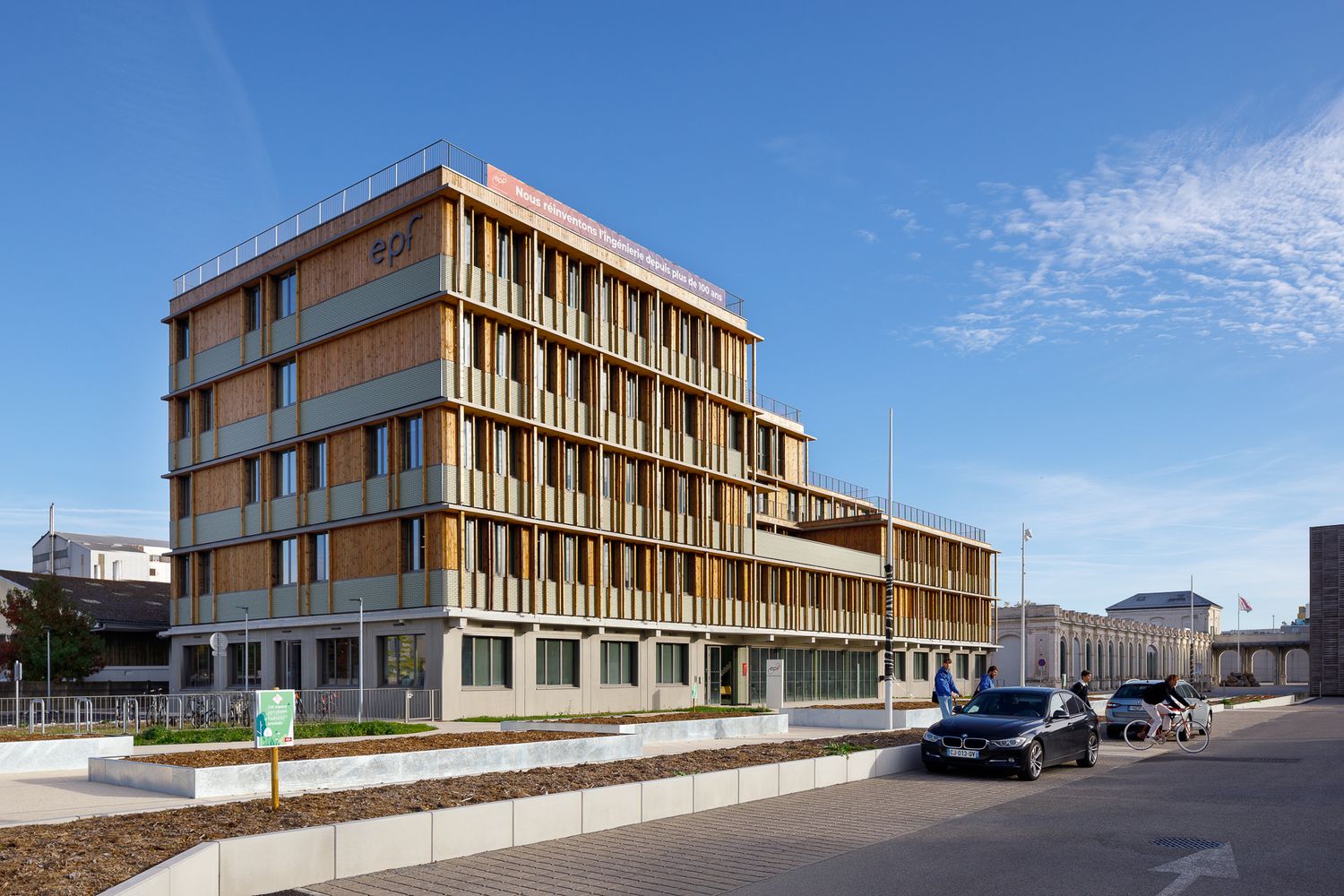 Immeuble de bureaux moderne de quatre étages, doté de brise-soleil en bois et d'un bardage végétalisé, situé à côté d'un parking urbain.