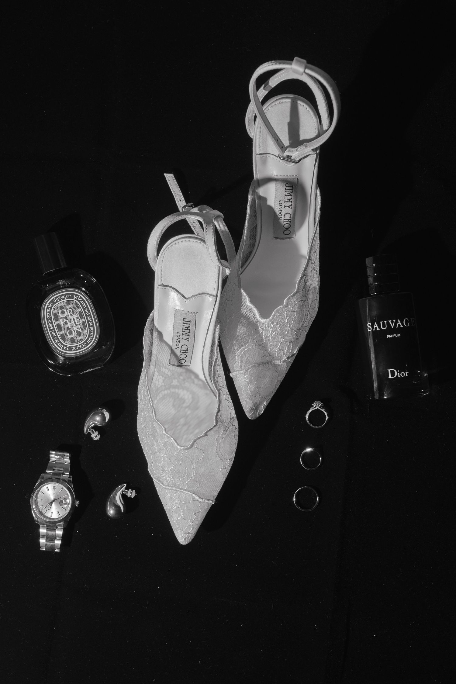 Black and white flat lay of Jimmy Choo lace heels, Dior Sauvage, a watch, rings, and earrings on dark background.