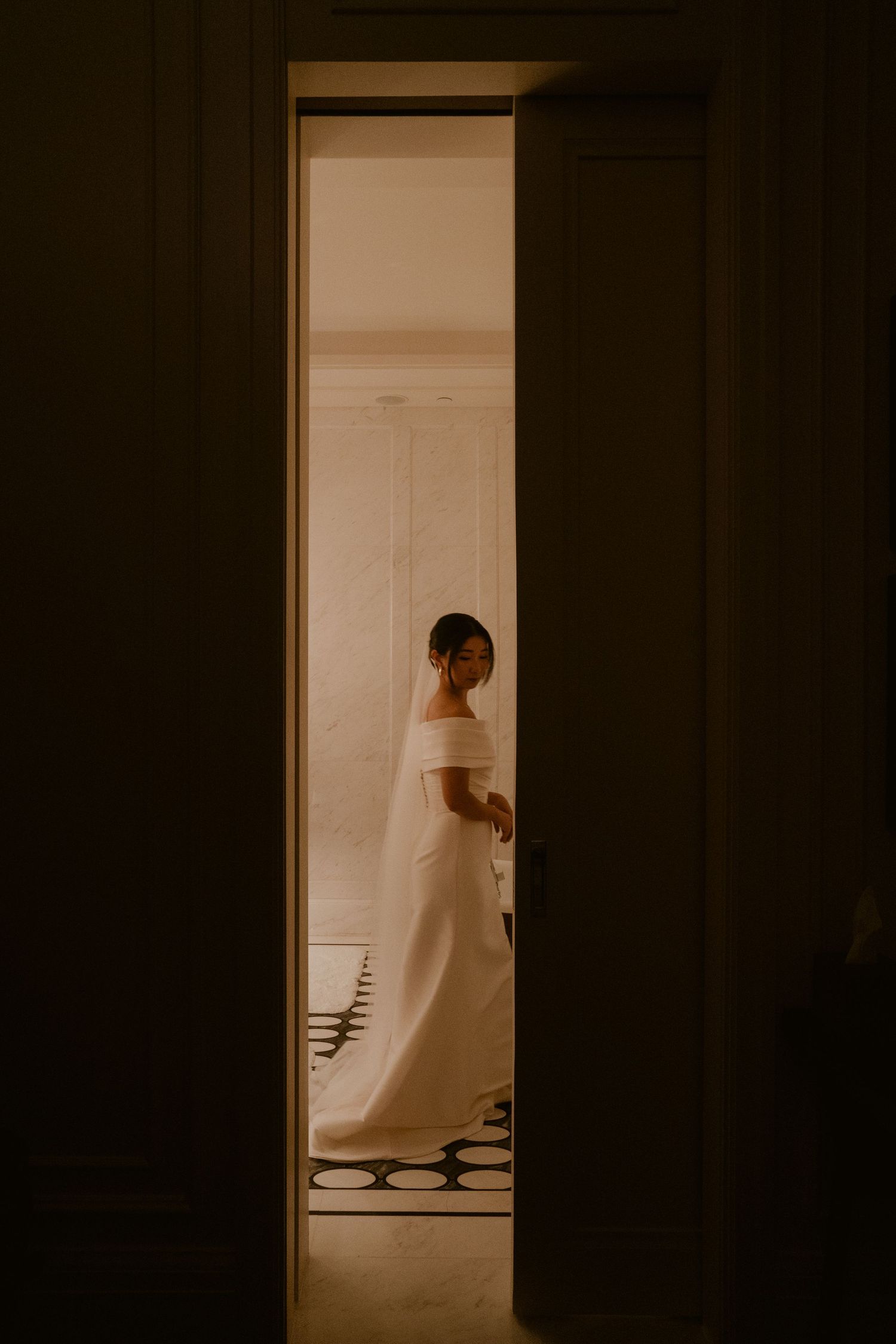 Bride in elegant off-shoulder white wedding gown and veil seen through a doorway in soft warm light.