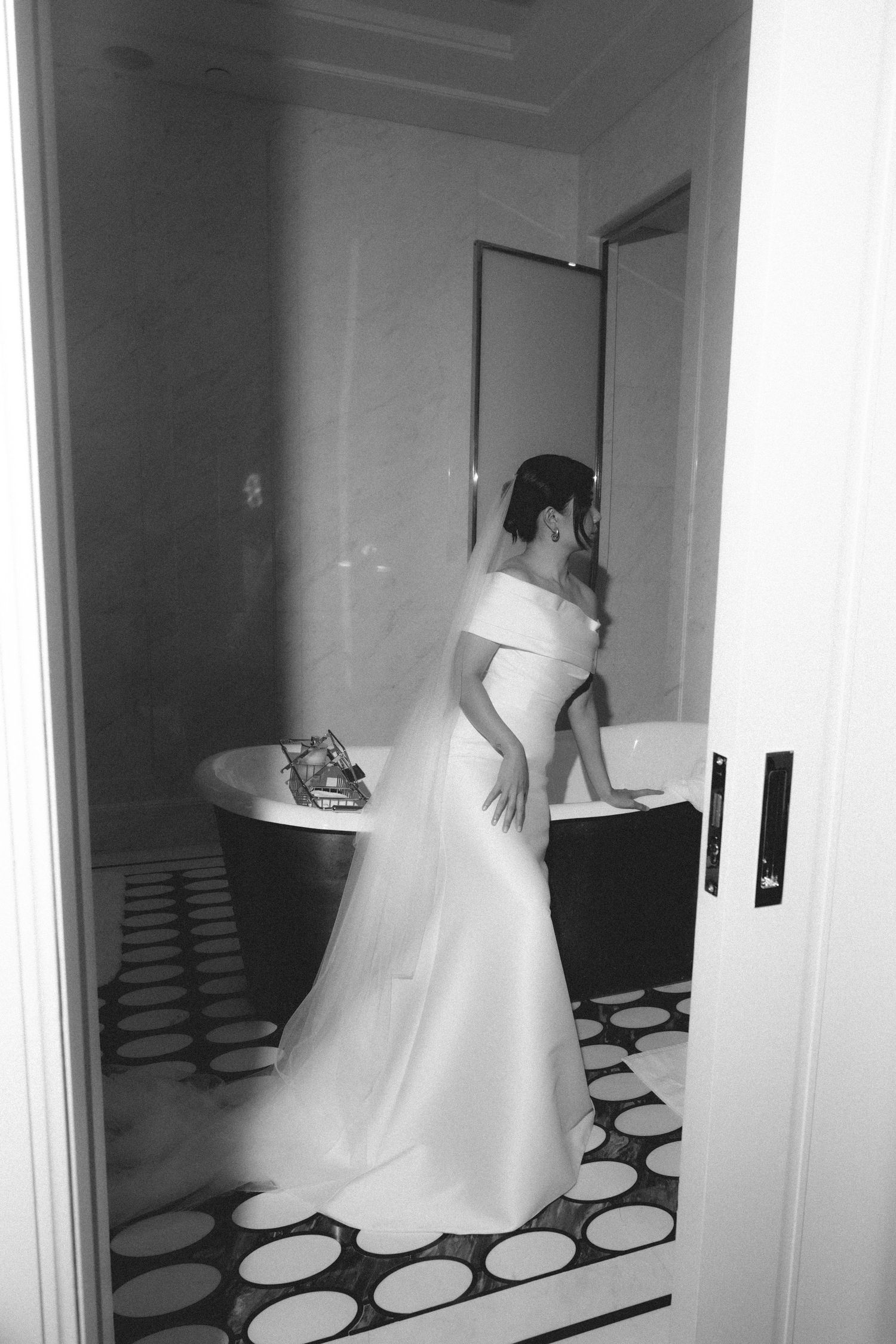 Bride in off-shoulder white wedding gown standing in luxurious black and white tiled bathroom.