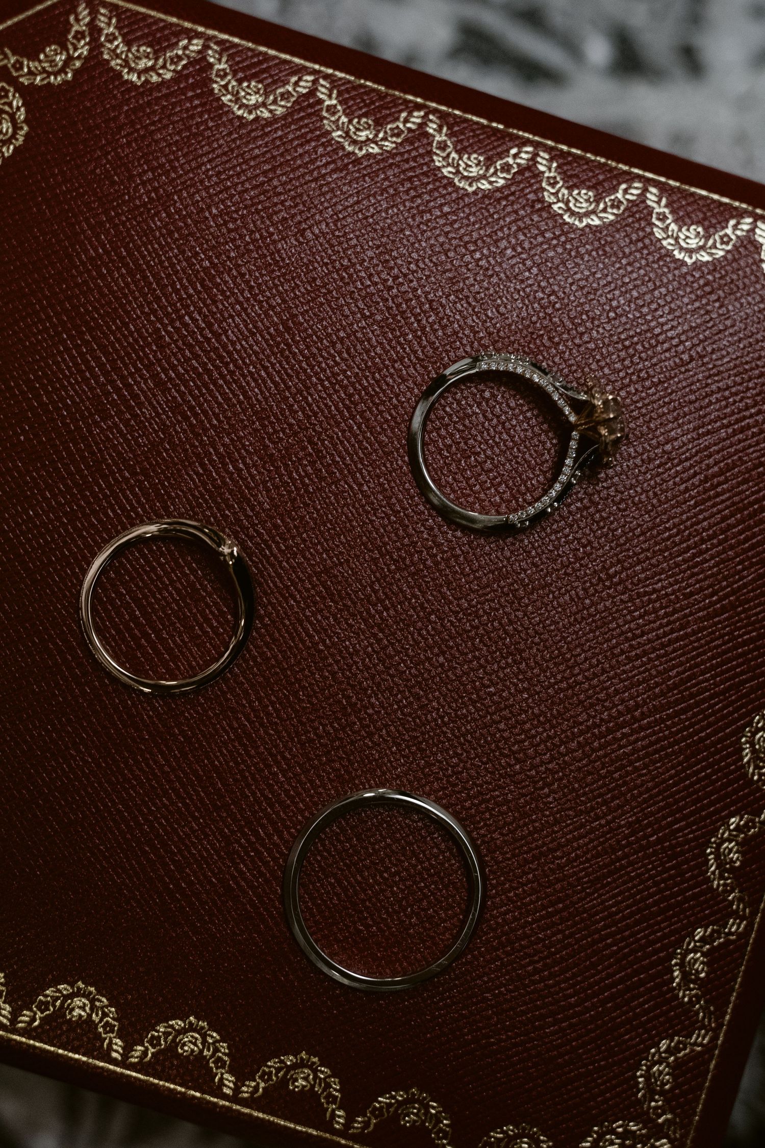 Three elegant rings displayed on a burgundy leather box with gold filigree border detailing.