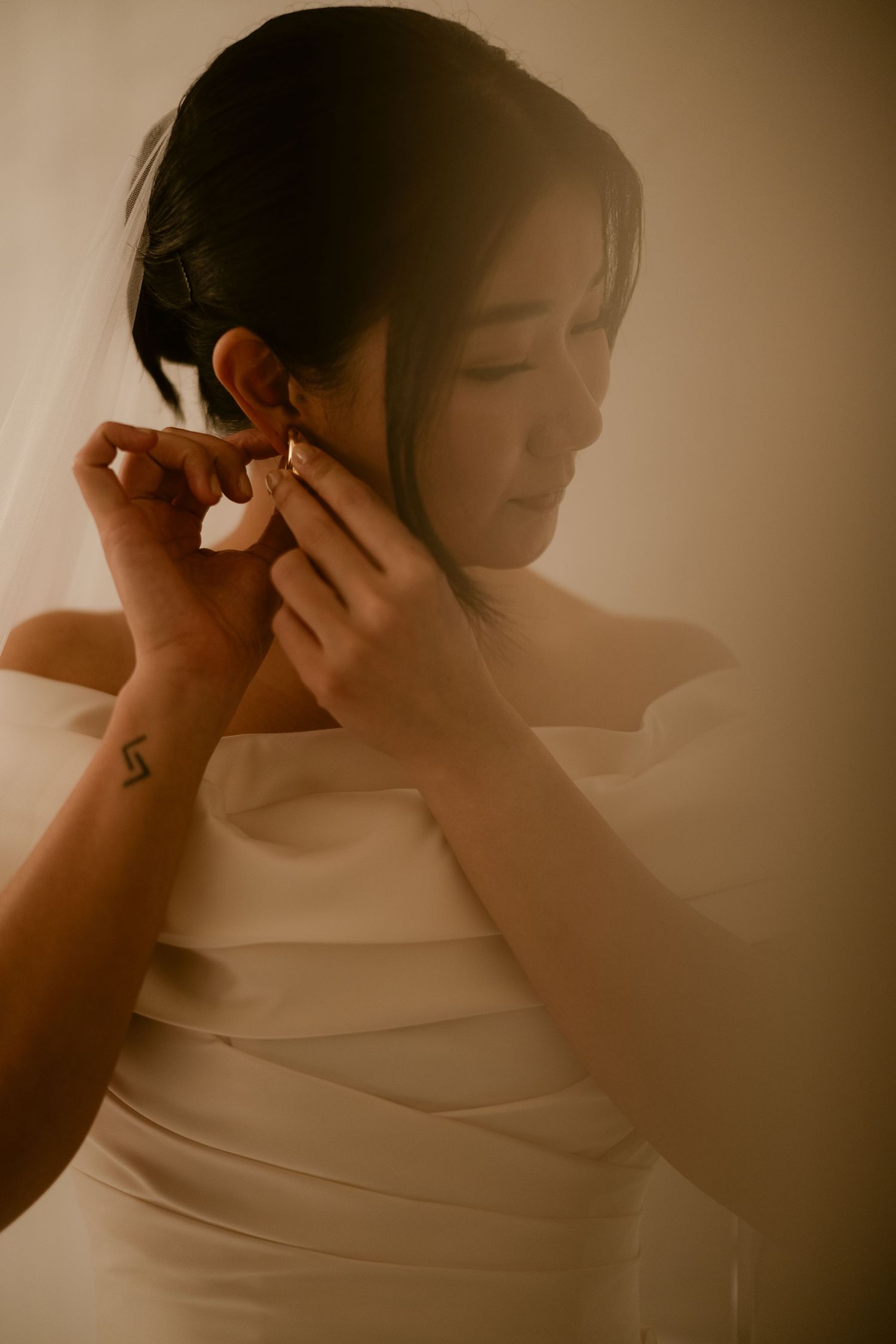 Bride in white strapless gown putting on gold earrings in warm golden light.