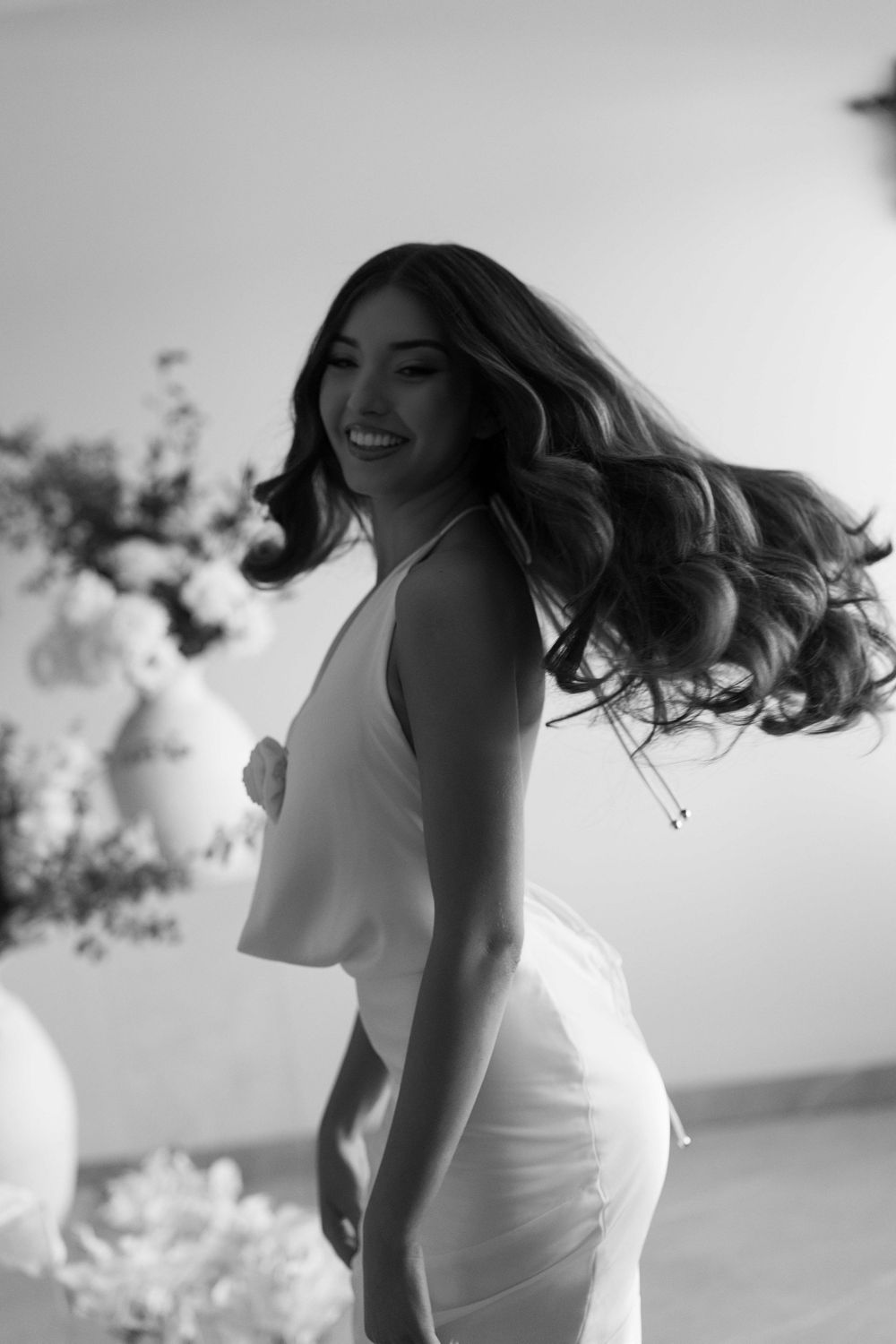 Black and white photo of a smiling woman in a white dress with flowing hair, posing elegantly indoors.