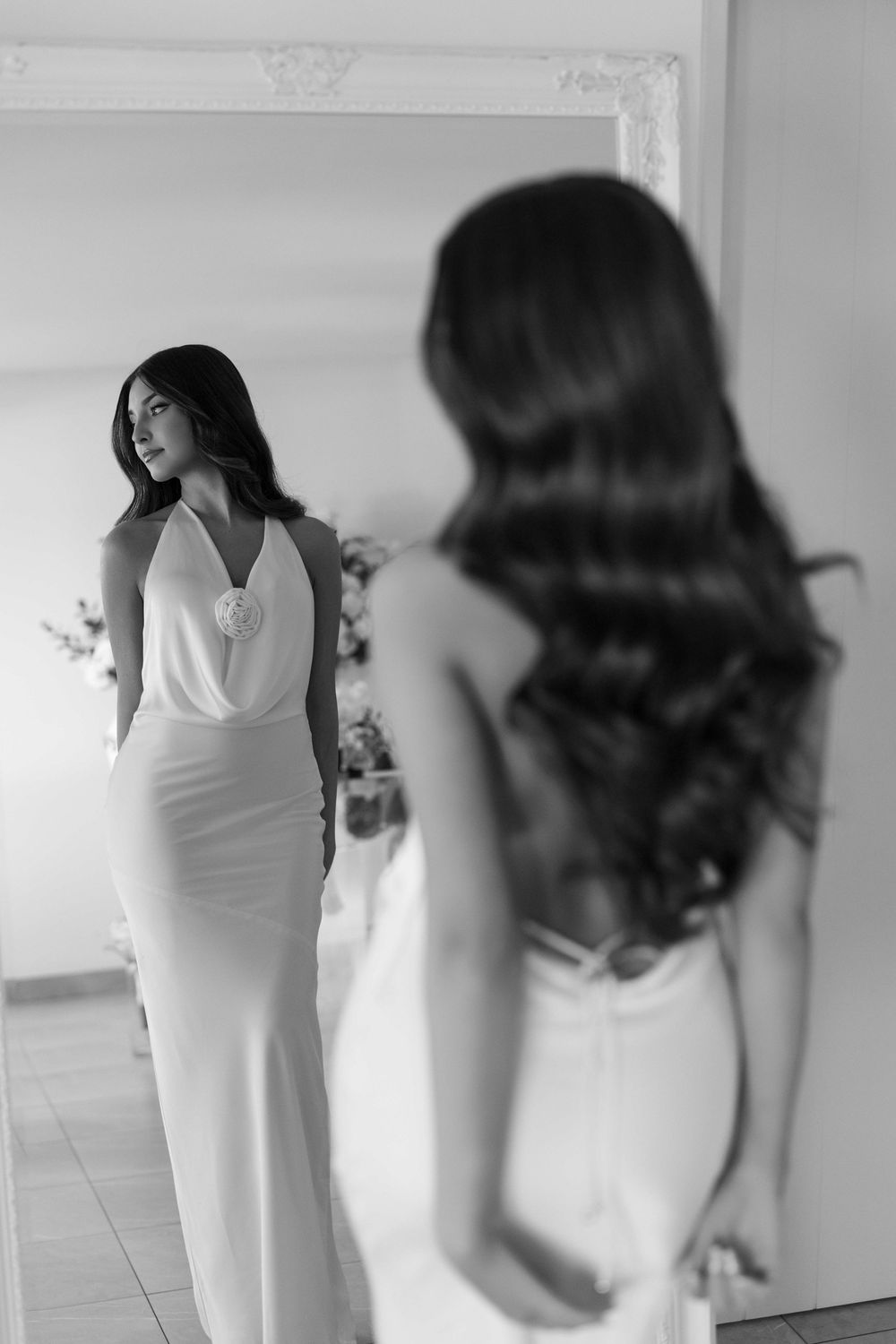 Black and white photo of a bride in a sleek white gown admiring her reflection in a large ornate mirror.