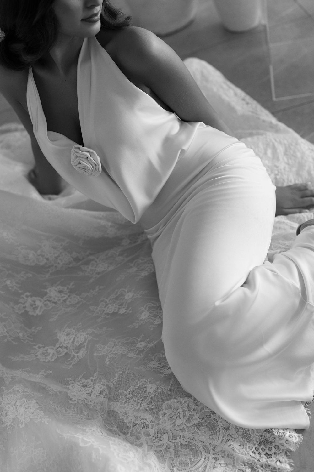 Black and white photo of a woman in an elegant white satin halter gown with floral detail, seated gracefully on lace fabric.