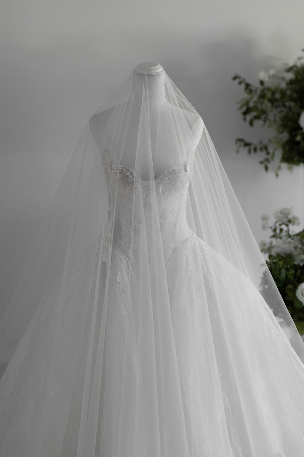 Elegant white bridal gown with lace bodice and flowing veil displayed on a mannequin against a soft gray background.