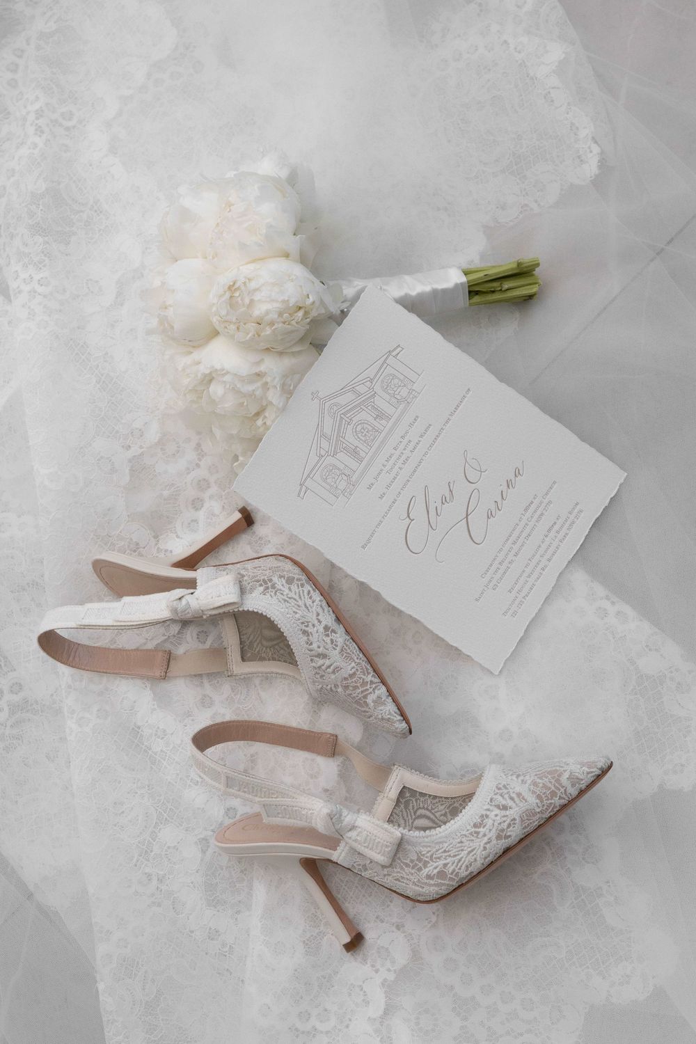 Elegant bridal flat lay with lace heels, white peony bouquet, and wedding invitation on soft tulle fabric.