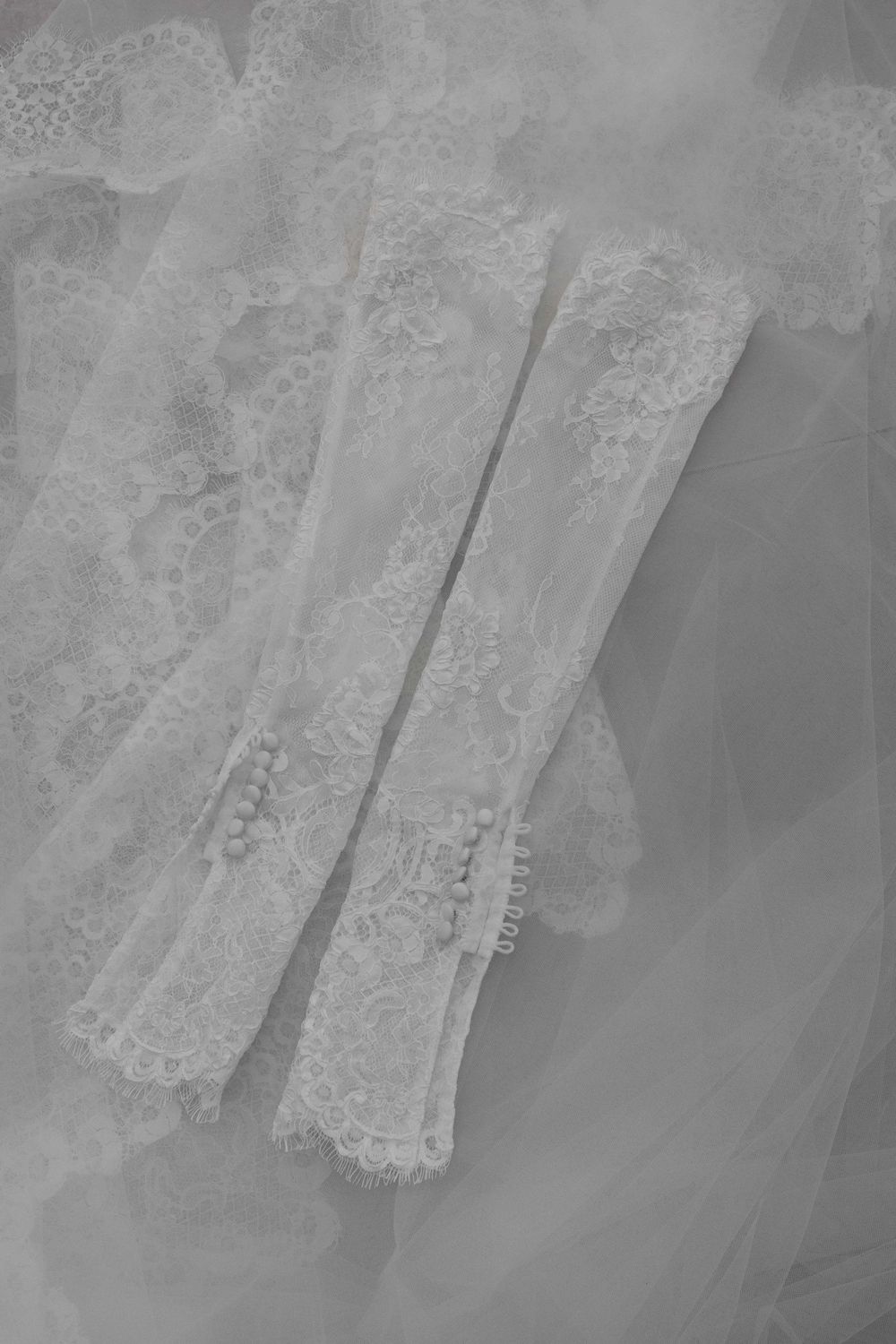 Elegant white lace bridal gloves with pearl buttons displayed on soft tulle fabric.