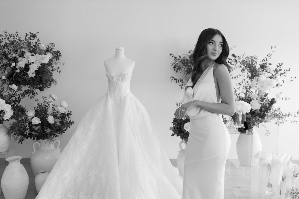 Black and white photo of a woman in a bridal boutique with a wedding gown on a mannequin and floral arrangements.
