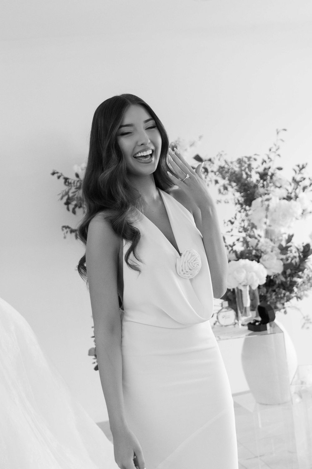 Smiling bride in elegant white halter gown with floral detail, laughing joyfully in black and white photo with floral arrangements.