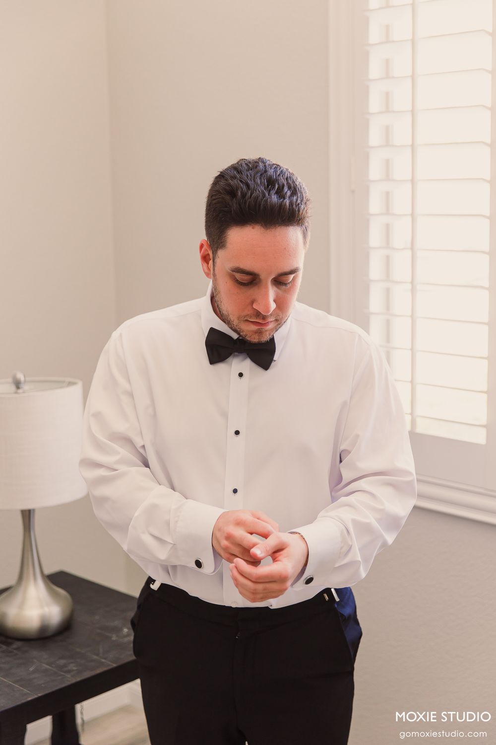 Groom in white tuxedo shirt and black bow tie adjusting cufflinks before wedding ceremony.