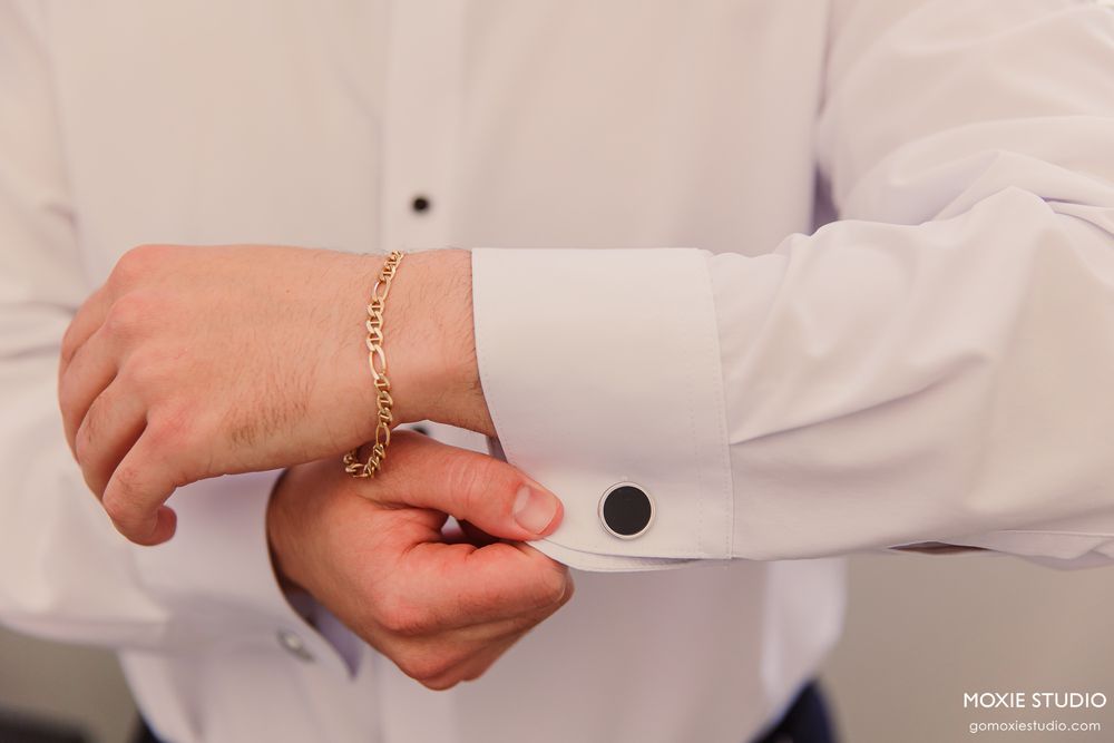 Man in white dress shirt fastening gold chain bracelet on wrist with black cufflink visible.