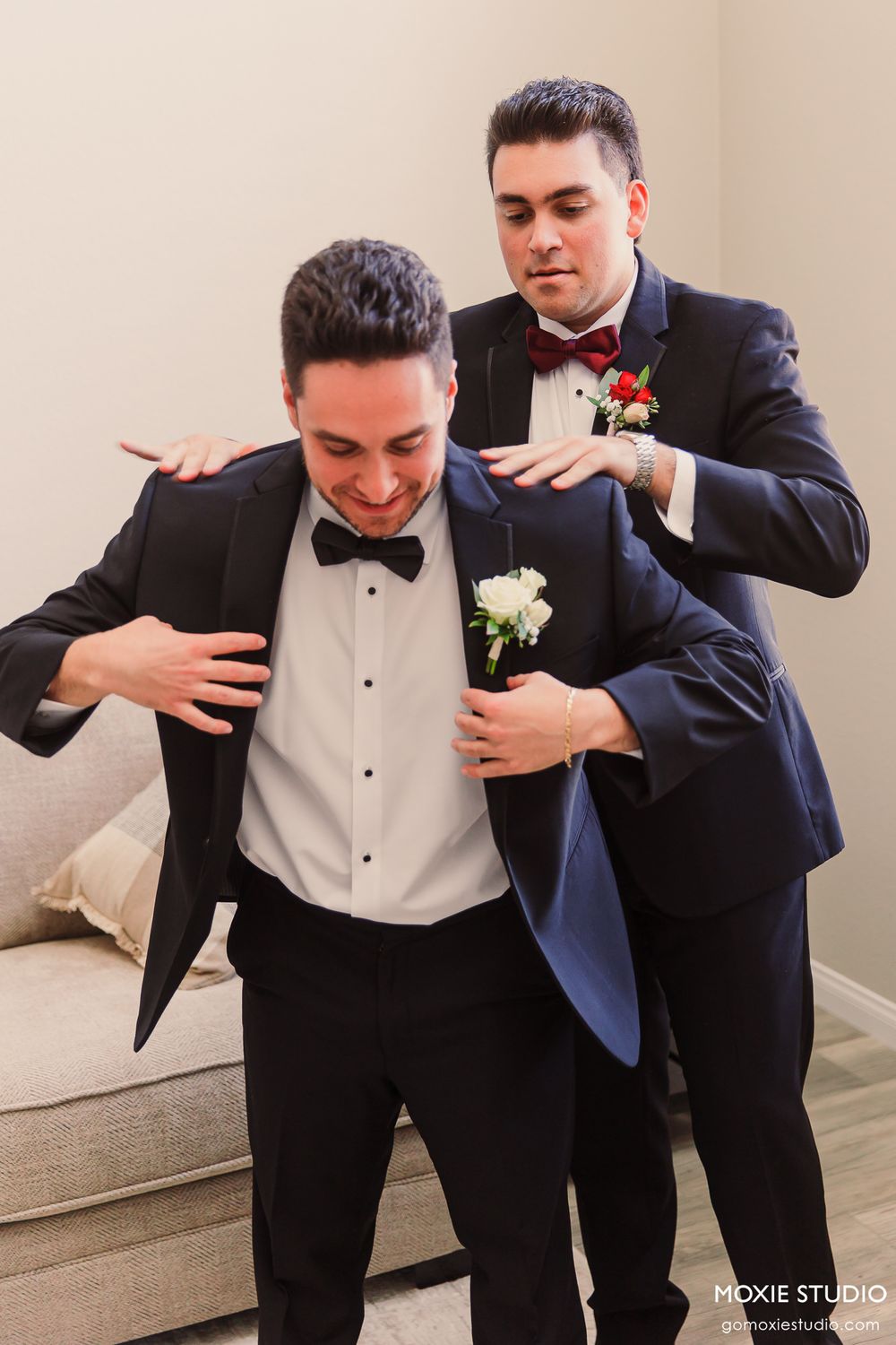 Groomsman helping groom adjust his black tuxedo jacket with boutonniere before wedding ceremony.