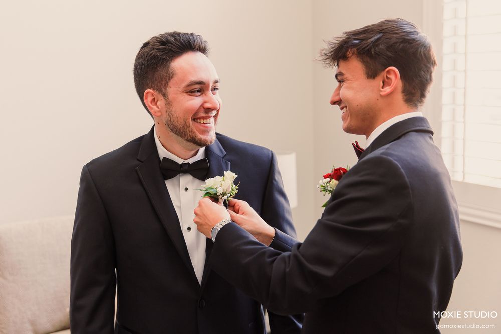 Groom smiling as best man pins white boutonniere on tuxedo lapel during wedding preparations.