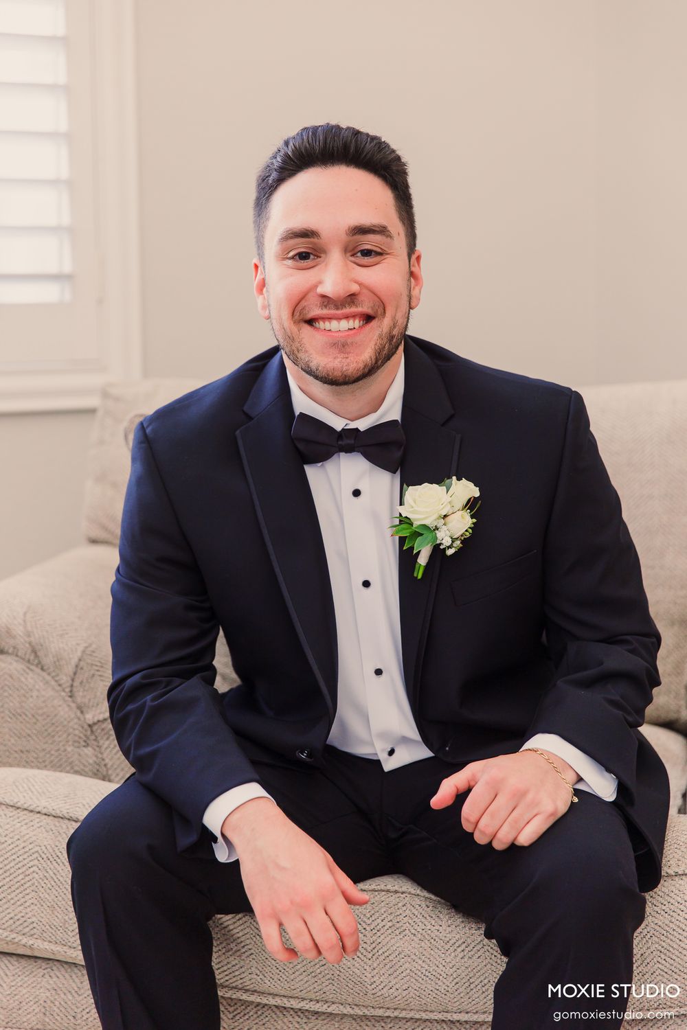 Smiling groom in navy tuxedo with white boutonniere sitting on couch before wedding.