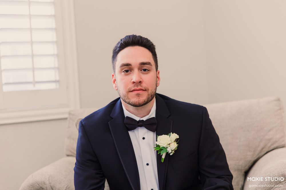Groom in navy tuxedo with black bow tie and white boutonniere sitting on couch before wedding.