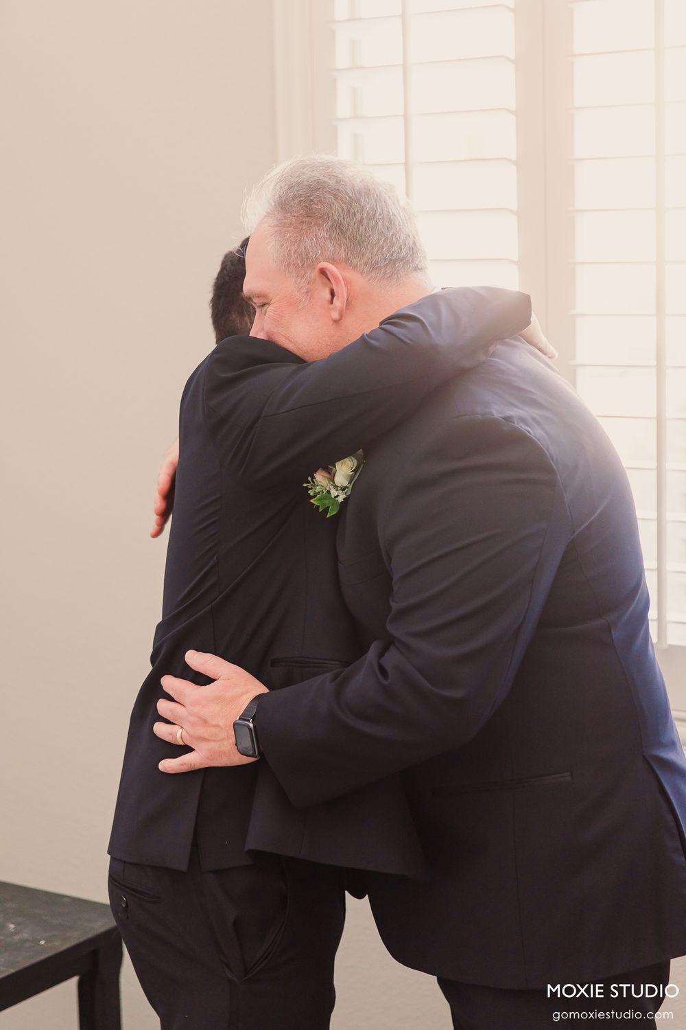Two men in navy suits share an emotional hug at a wedding, one wearing a white boutonniere corsage.