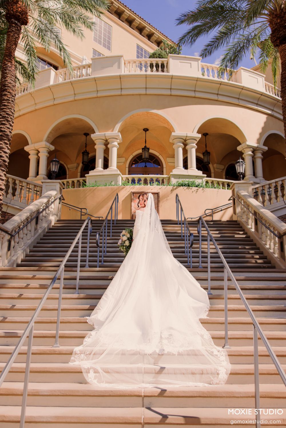 Bride in white gown with long veil ascending grand staircase at elegant venue.