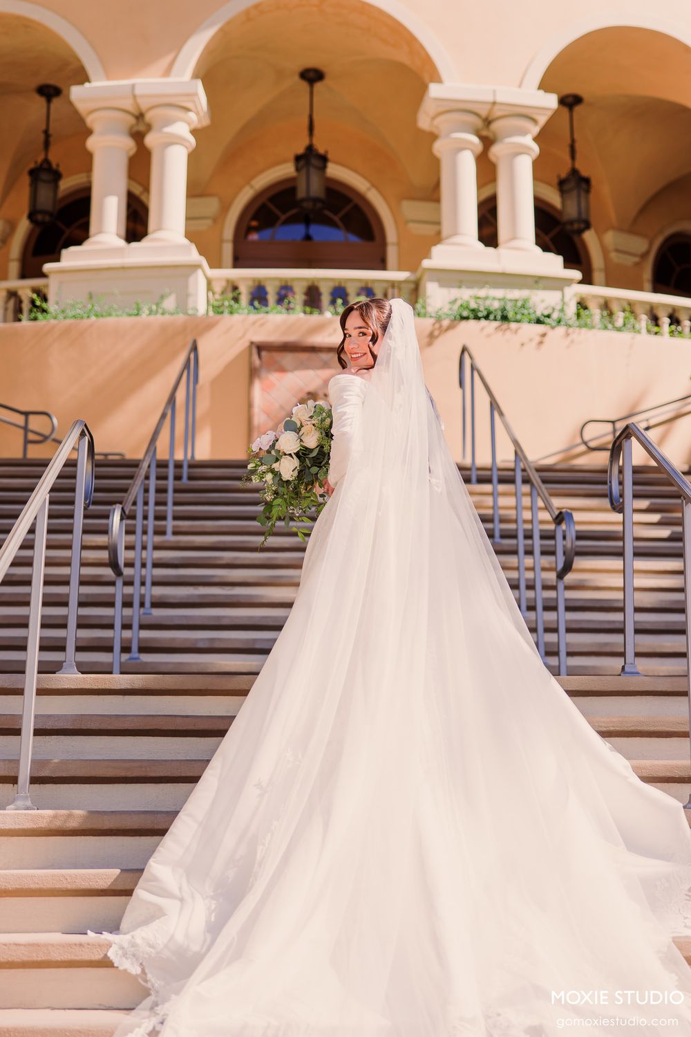 Bride in elegant white gown with long veil holds bouquet on grand staircase of ornate venue.