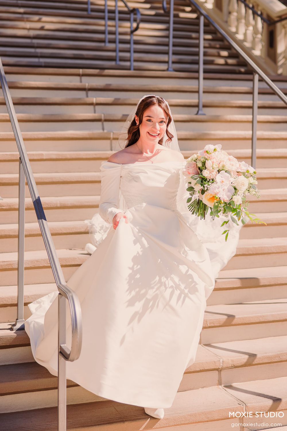 Bride in elegant off-shoulder white gown holding pastel bouquet on grand stone staircase.