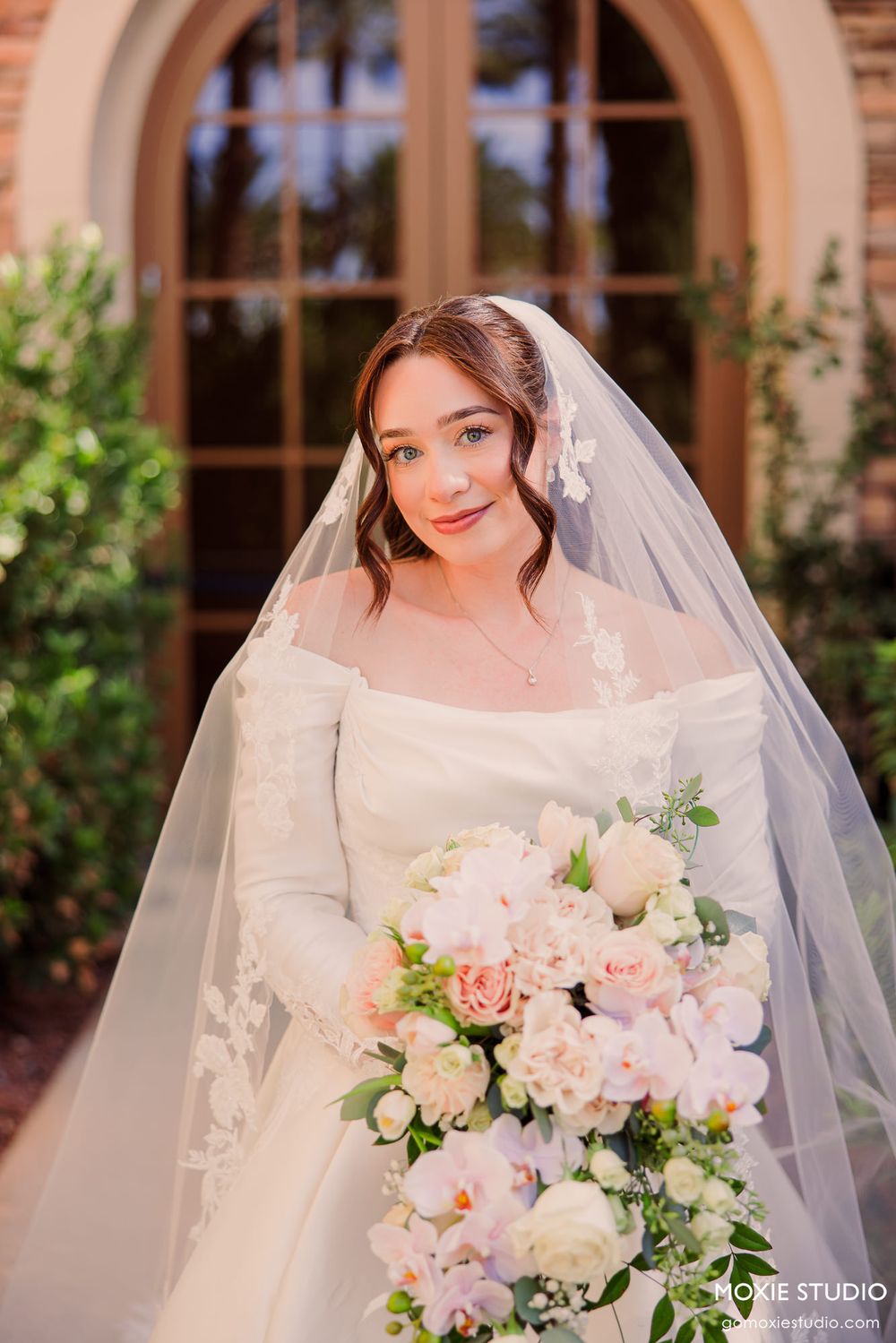Bride in white off-shoulder gown with lace veil holds pink floral bouquet outdoors at elegant venue.