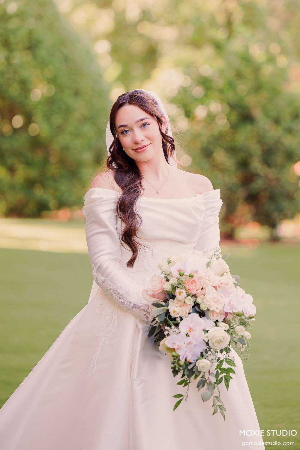 Elegant bride in off-shoulder white gown holds a soft pink and white floral bouquet in a lush garden setting.