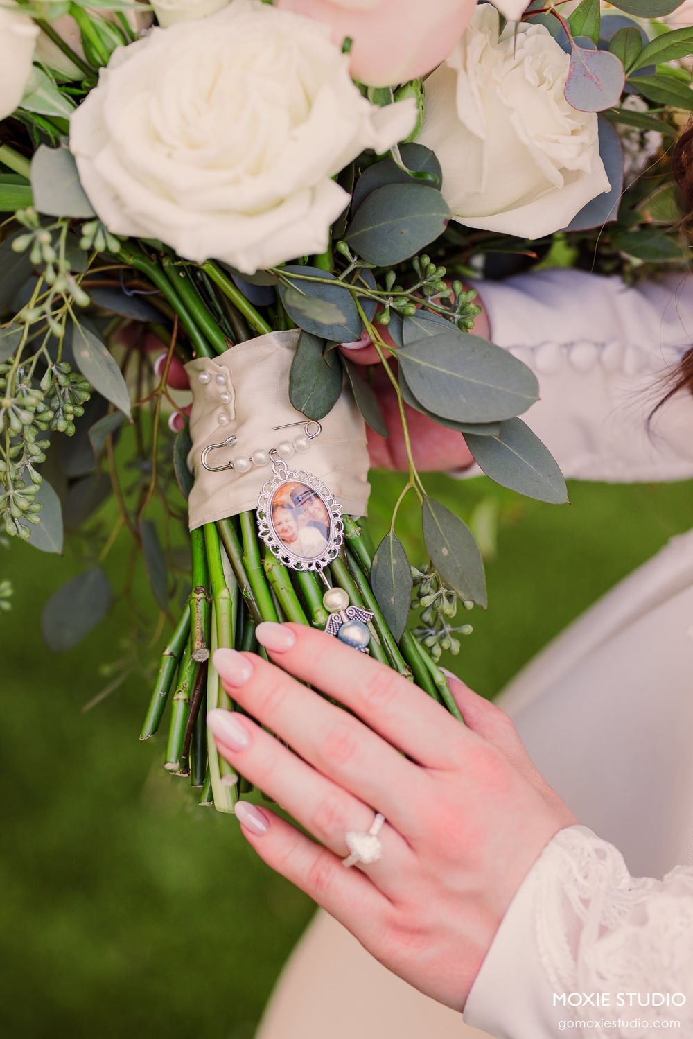 Bride holding a white rose bouquet with eucalyptus, a memorial photo charm, and wearing a diamond ring on her finger.