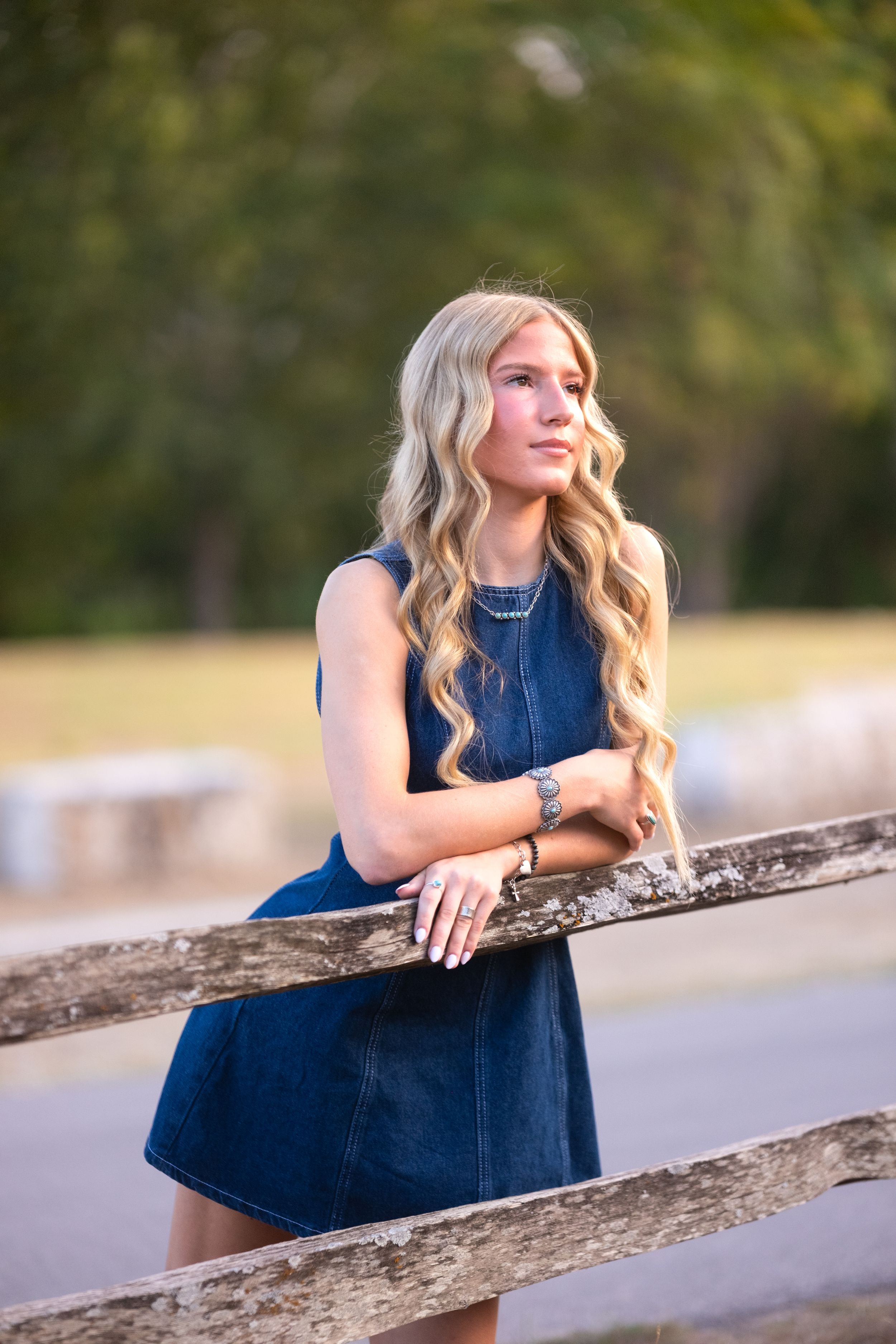 Blonde teenage girl in denim dress leans on rustic wooden fence during golden hour outdoors in Salado Creek, Salado Texas