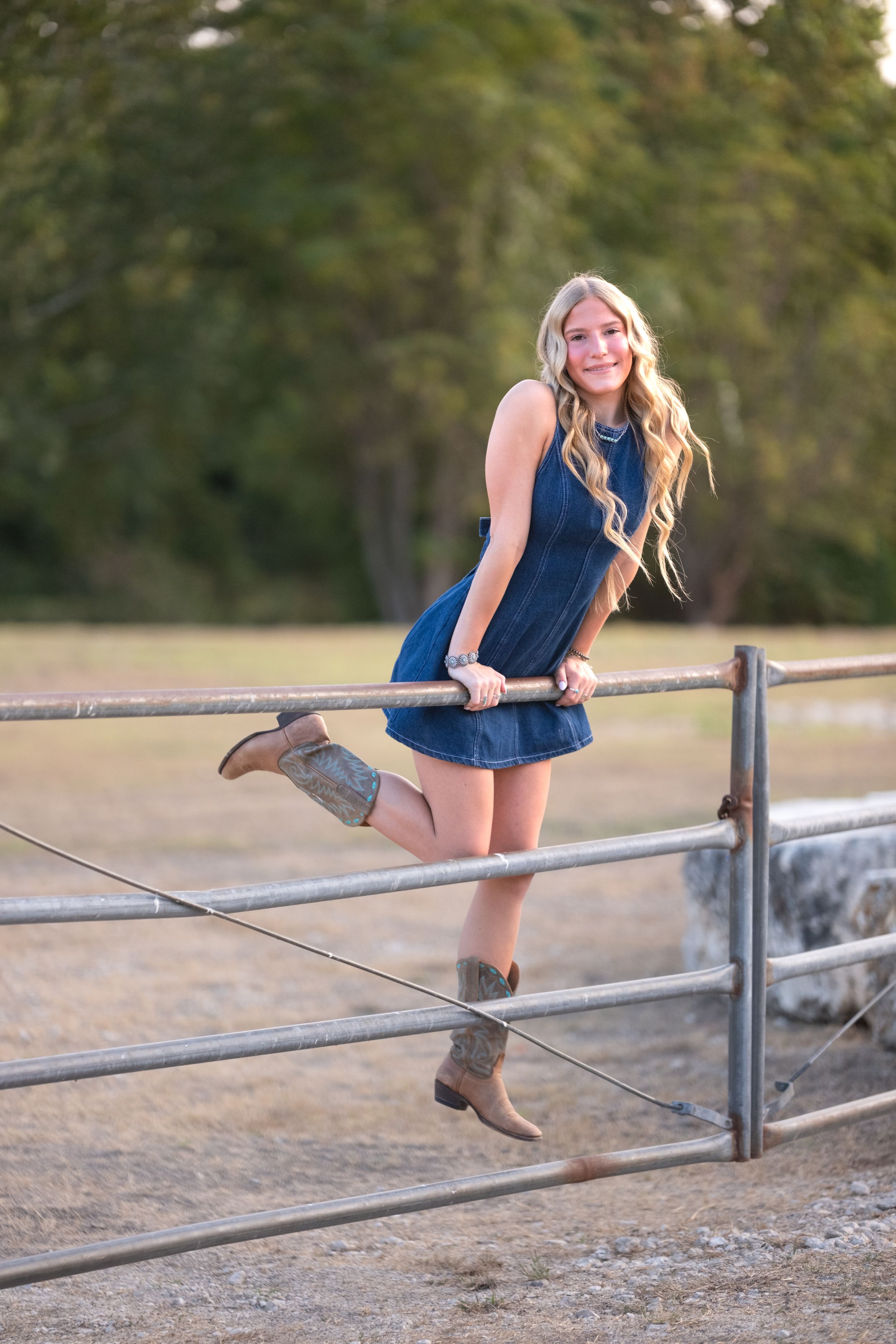 Smiling blonde teen in denim dress and cowboy boots posing on a metal farm gate outdoors in Salado Creek, Salado Texas