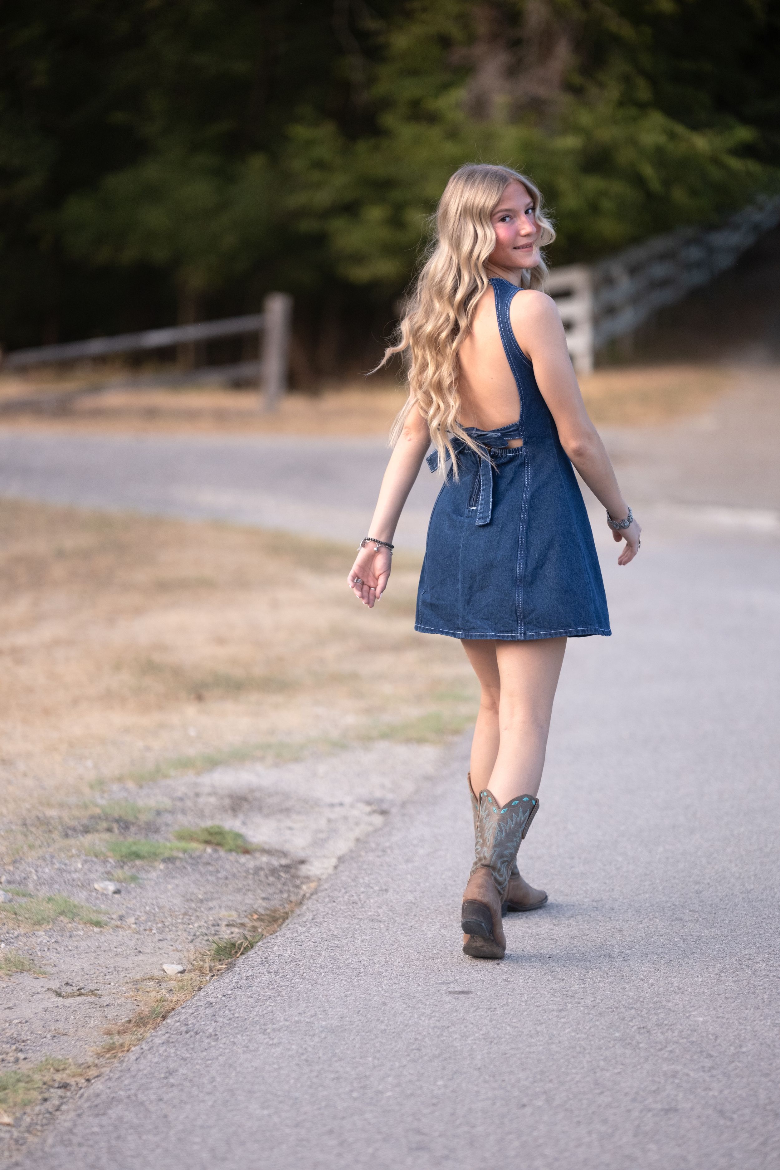 Blonde teen in backless denim dress and cowboy boots walking along a rural road at sunset in Salado Creek, Salado Texas