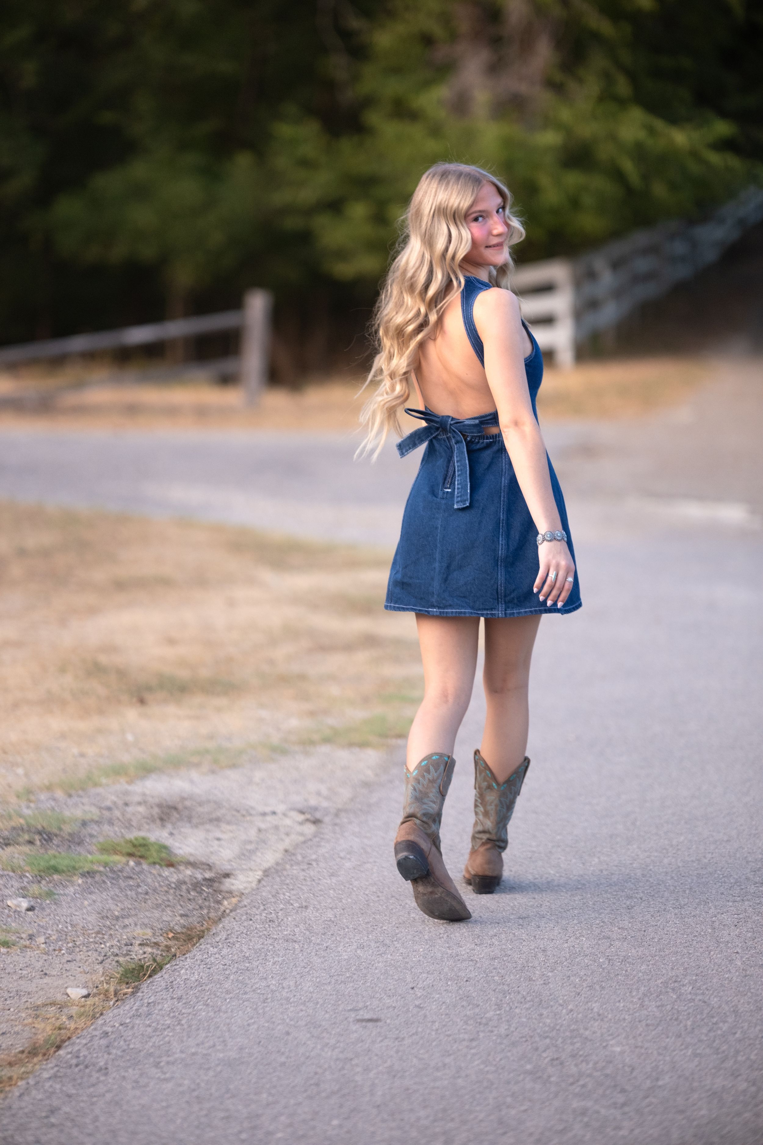 Blonde teen in open-back denim dress and cowboy boots posing on a country road at golden hour in Salado Creek, Salado Texas