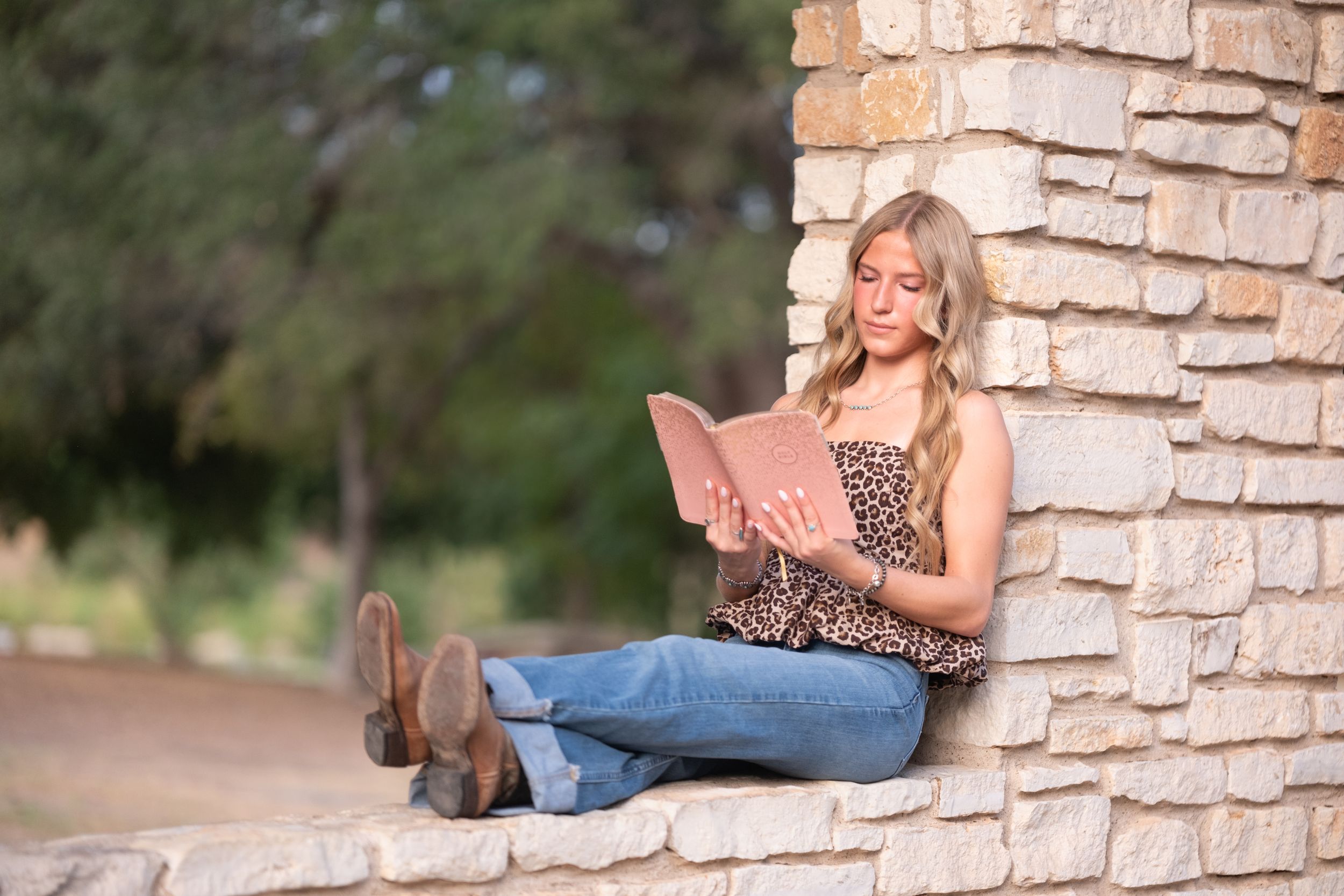 Blonde teen girl sitting against a stone pillar reading a book outdoors in natural light in Salado Creek, Salado Texas