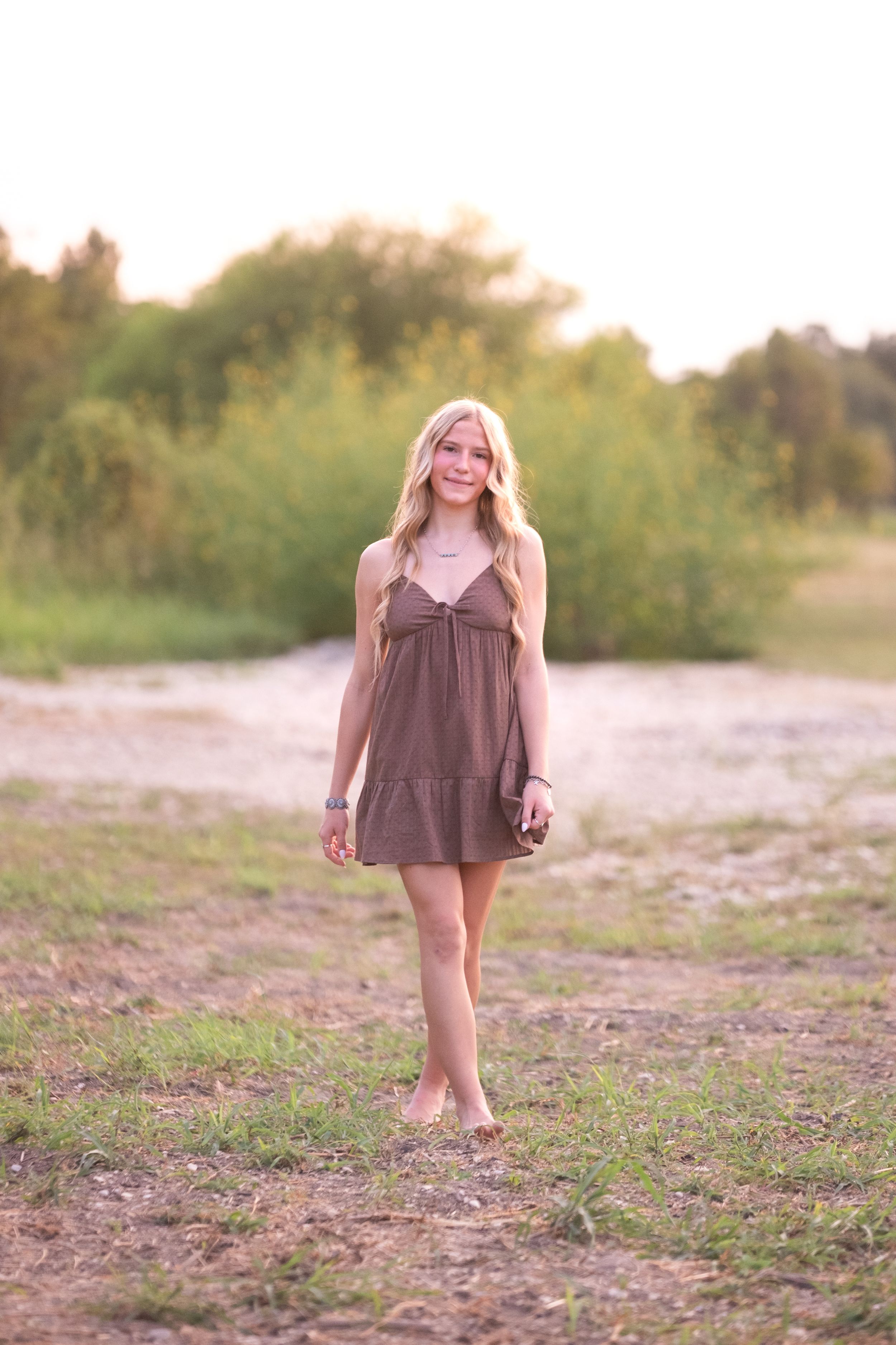 Smiling blonde teen in a brown sundress walking barefoot through a grassy field at sunset in Salado Creek, Salado Texas