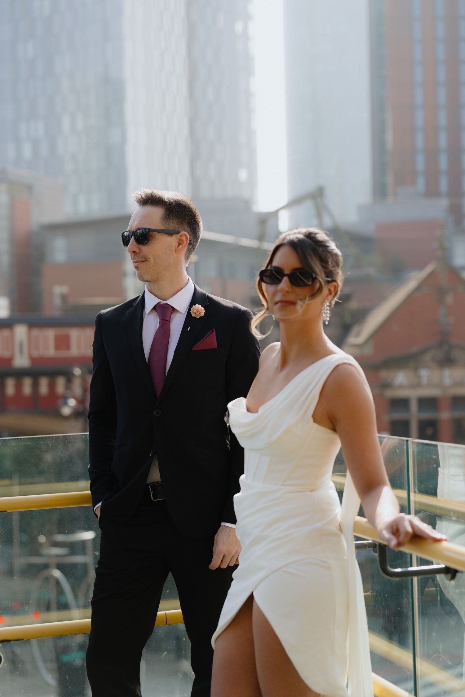 Stylish bride in white asymmetric dress and groom in black suit with burgundy tie pose on rooftop terrace.